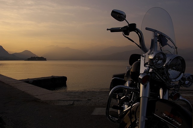Une Harley-Davidson avec une bulle moto fumée est garée au bord d'un lac au coucher du soleil, avec des montagnes en arrière-plan.