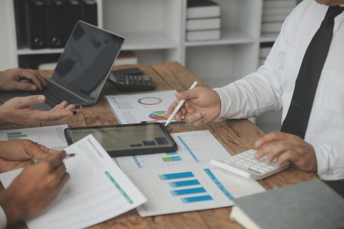Une équipe de professionnels en pleine réunion de travail analysant des graphiques financiers et des rapports de gestion d'actifs sur une table en bois.