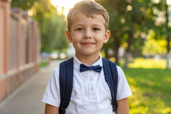 Un jeune garçon souriant en polo blanc et nœud papillon porte un sac à dos bleu foncé.
