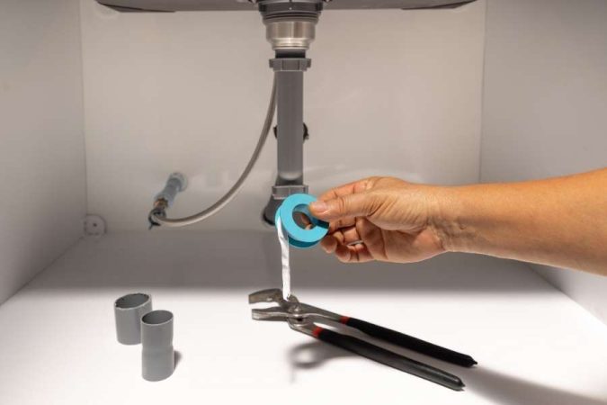 Une main tenant un rouleau de ruban d'étanchéité bleu, avec des outils et un raccord de tuyau gris posés pour une installation sous un évier.