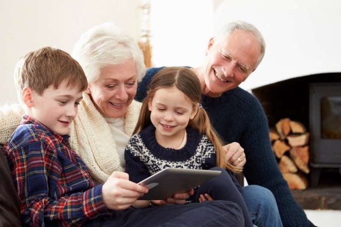 Un grand-père et une grand-mère souriants partagent un moment complice autour d'une tablette numérique avec leurs petits-enfants.